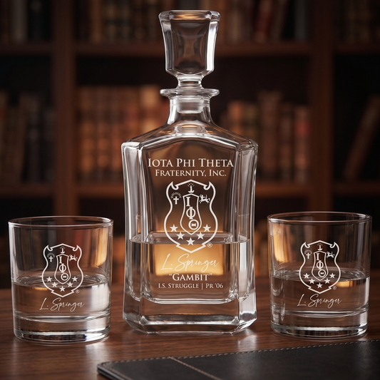A clear glass decanter and two matching lowball glasses engraved with an Iota Phi Theta Fraternity crest and the name L. Spiga, displayed on a wooden table with a blurred bookshelf background.